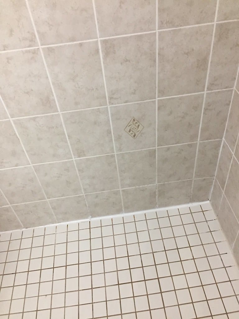 A tiled shower corner with clean white grout lines between the pale wall tiles and brown-stained grout between the small floor tiles below.