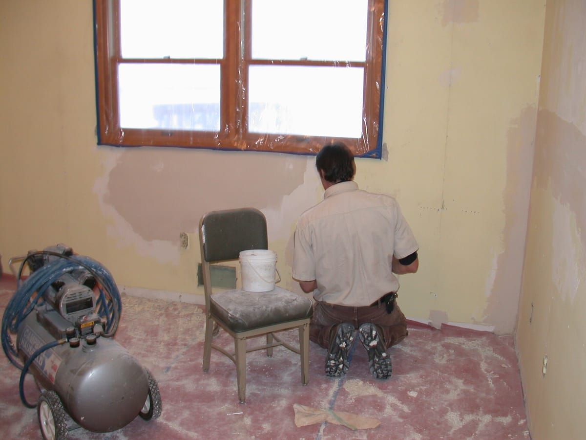A decorator kneeling at a bedroom wall, smoothing joint compound over a repair, with pale filled patches feathered across the wall above and dust across the floor.