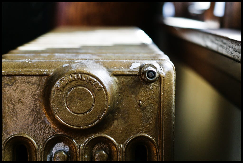 The top corner of an ornate cast iron column radiator, its bronze paint catching light from a window behind it.