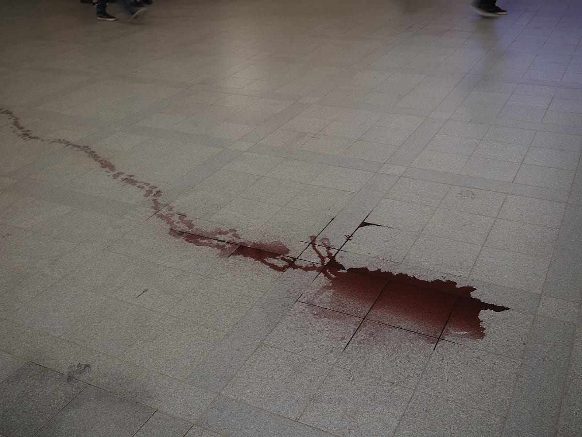 A trail of spilled red wine pooling across grey floor tiles, still wet, with the colour darkest where it has collected