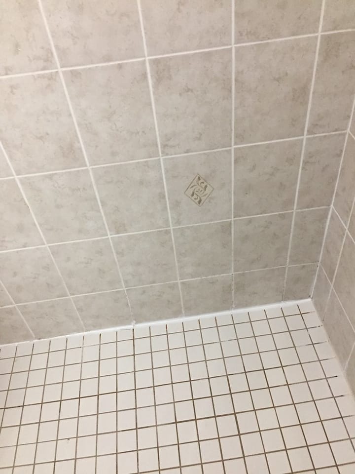 A tiled shower corner with clean white grout lines between the pale wall tiles and brown-stained grout between the small floor tiles below.