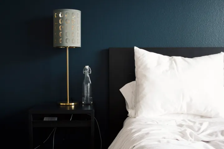 A white pillow and white bed sheets on a made bed against a dark blue wall, with a lamp on the bedside table