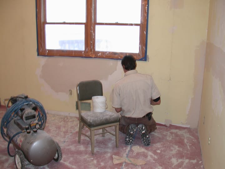 A decorator kneeling at a bedroom wall, smoothing joint compound over a repair, with pale filled patches feathered across the wall above and dust across the floor.