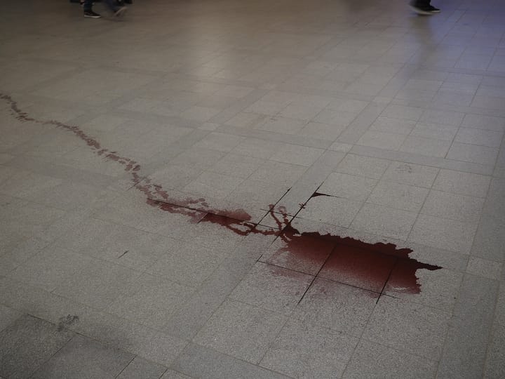 A trail of spilled red wine pooling across grey floor tiles, still wet, with the colour darkest where it has collected