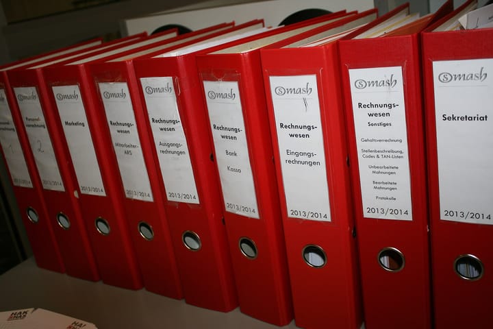 A row of red lever-arch binders on a shelf, each spine labelled by subject and by year.