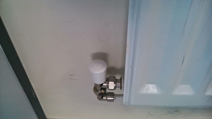 The white-capped lockshield valve and chrome pipe union at the bottom corner of a white panel radiator, against a pale wall.