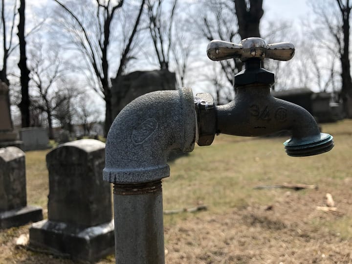 A standard outdoor hose spigot with a cross-shaped metal handle and a threaded hose end, on a galvanized standpipe, with bare winter trees and headstones blurred in the background.